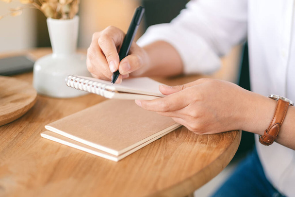 Imagen de cerca de una mujer sosteniendo y escribiendo en un cuaderno en blanco sobre una mesa de madera  - Foto, imagen