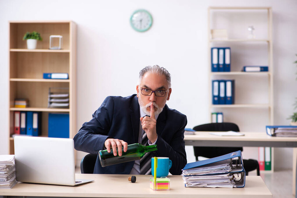 Old male employee drinking alcohol in the office - 写真・画像