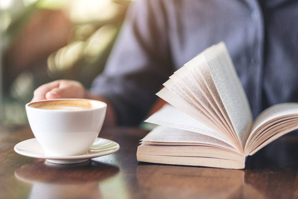 Nahaufnahme einer Frau, die ein Buch hält und liest, während sie Kaffee auf einem Holztisch trinkt - Foto, Bild