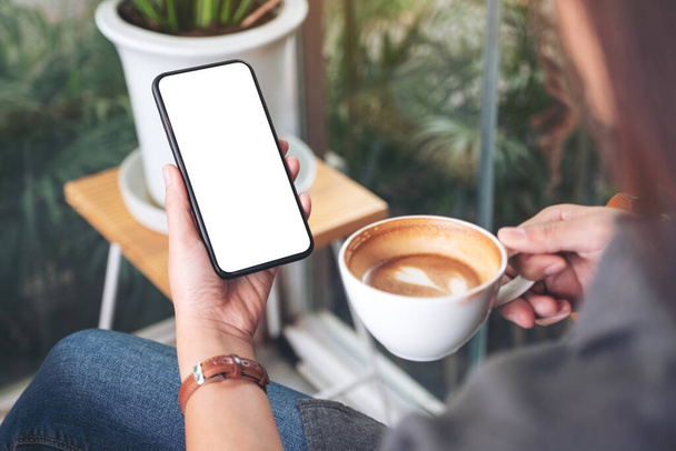 Mockup image of a woman holding and using black mobile phone with blank desktop screen while drinking coffee in cafe - Fotografie, Obrázek