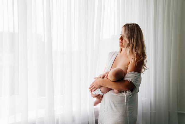 mom feeds baby standing at the window. breast-feeding. - Фото, изображение