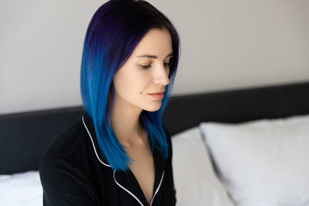 Beautiful woman with blue hair in black nightwear sitting on bed after sleep - Photo, Image