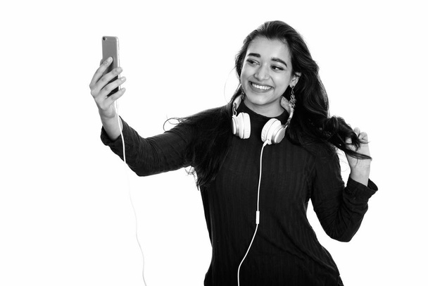 Young happy Indian woman smiling while wearing headphones around neck and taking selfie picture with mobile phone - Foto, Bild