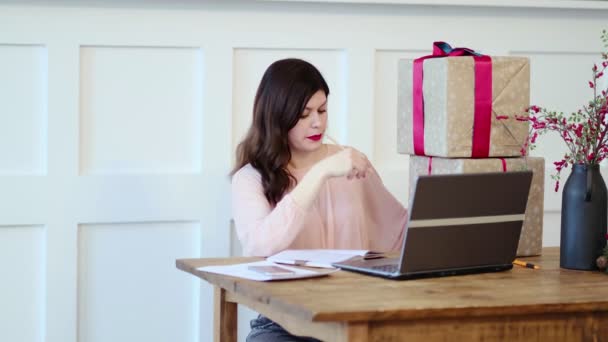 Pretty young lady sitting at desk and typing on laptop and talking with present boxes icons. box receive good online shop purchase at home, post mail shipping delivery concept - Footage, Video