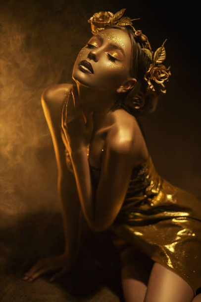 Portrait Beauty fantasy woman autumn queen, face in gold paint golden shiny skin. Fashion model girl posing. Black studio. Glamorous crown, wreath roses flowers, jewellery accessories, metallic makeup - Photo, Image