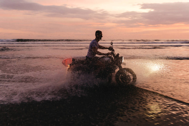 Surfer rides on motorbike with surfboard at sunset ocean beach. - Fotó, kép