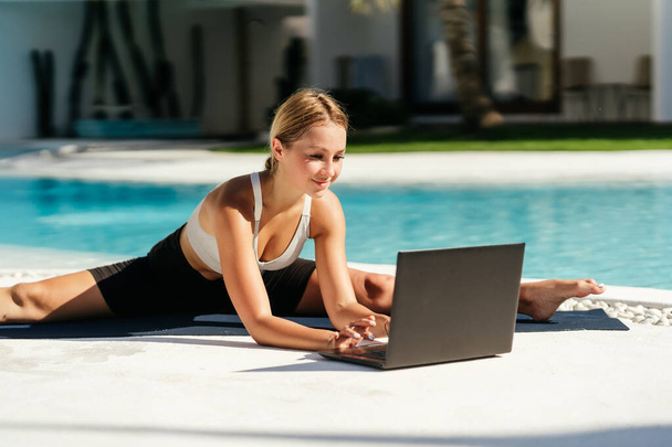 Young fit woman doing yoga stretching exercise outdoor near swimming pool in front of laptop. - Foto, Imagen
