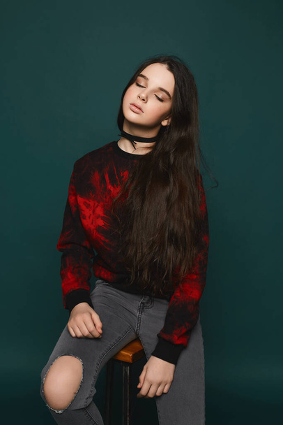 Beautiful teenage model girl in stylish black and red sweatshirt and a black choker on her neck posing at the dark-green background - Фото, зображення