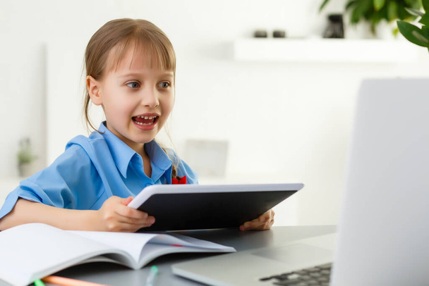 Милая маленькая девочка сидит за столом со своим ноутбуком и учится в Интернете - Фото, изображение