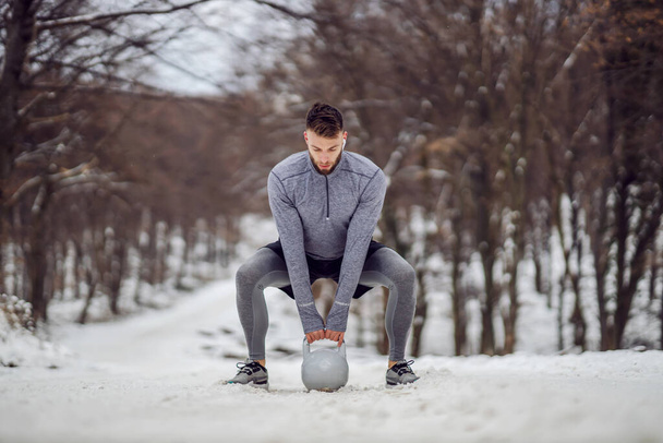 Bodybuilder tillen kettlebell terwijl hurken in het bos op besneeuwde parcours in de winter. Bodybuilding, Wintersport, Fitness - Foto, afbeelding