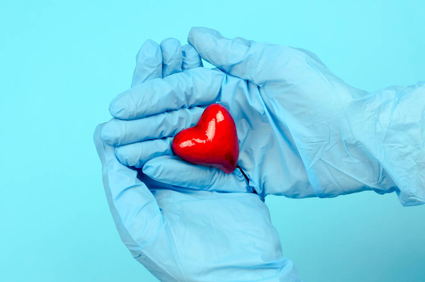 Ręce w niebieskich rękawiczkach medycznych trzymają serce na niebieskim tle. Koncepcja opieki zdrowotnej. Światowy Dzień Serca - Zdjęcie, obraz