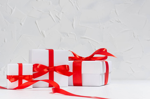 Gentle white gift boxes with red silk bow on wood table in white interior as festive anniversary background. - Fotoğraf, Görsel