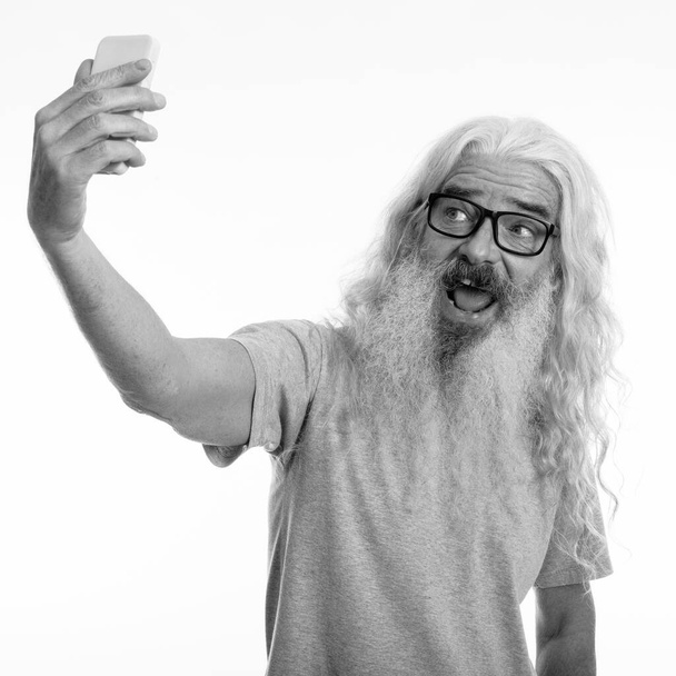 Studio shot of happy senior bearded man smiling while taking selfie picture with mobile phone - Photo, Image