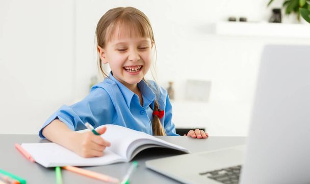 Az okos kislány elvégzi a házi feladatot a nappaliban. Az íróasztalánál ül, tollal ír a tankönyvében, és laptopot használ.. - Fotó, kép