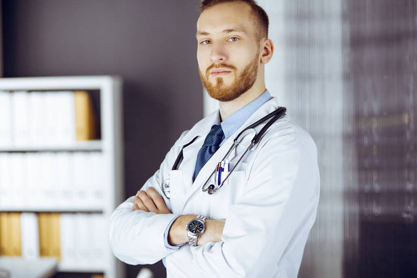 Red-bearded doctor standing straight in clinic near his working place. Portrait of physician. Medicine and healthcare concept - Zdjęcie, obraz