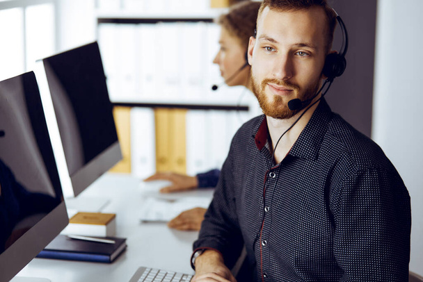 Red-bearded businessman talking by headset near his female colleague while sitting in modern sunny office. Diverse people group in call center - Foto, Bild