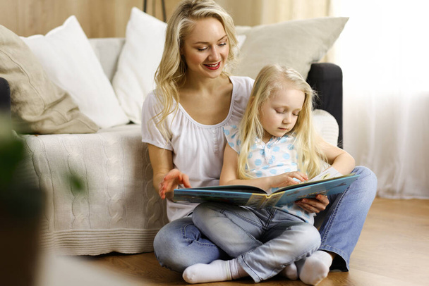 Happy family. Blonde young mother reading a book to her cute daughter while sitting at wooden floor. Motherhood concept - Foto, Bild