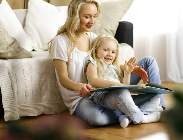 Happy family. Blonde young mother reading a book to her cute daughter while sitting at wooden floor. Motherhood concept - Zdjęcie, obraz