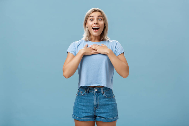 Porträt einer verblüfften und charmanten attraktiven blonden Studentin mit gebräunter Haut in Jeanshosen und Sommer-T-Shirt, die keuchend die Handflächen auf der Brust hält und freudig dankbar und zufrieden lächelt - Foto, Bild
