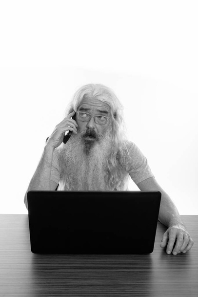 Plan studio d'un homme barbu âgé pensant et parlant sur un téléphone portable tout en utilisant un ordinateur portable sur une table en bois - Photo, image