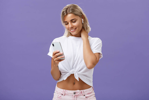 Woman receiving warm romantic message being delighted and touched feeling love gazing at smartphone screen wearing wireless earphones touching neck gently being sensitive and feminine - Photo, Image