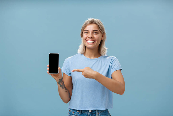 Cute enrgized girl showing smartphone. Charming blonde young female with cool tattoos laughing and smiling from happiness and joy pointing at device holding phone with screen faced to camera - Foto, Imagen