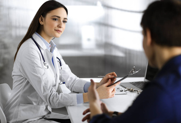Friendly young woman-doctor is listening to her patient, while they are sitting together at the desk in a cabinet. Physician is holding a clipboard in her hands for filling up medication - Photo, image