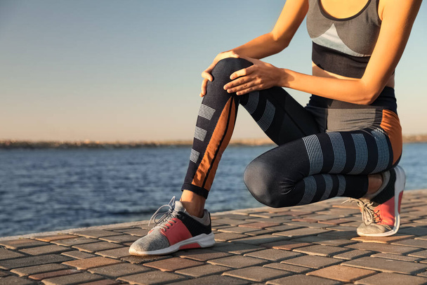 Young woman in sportswear having knee problems near river at sunset, closeup - Zdjęcie, obraz