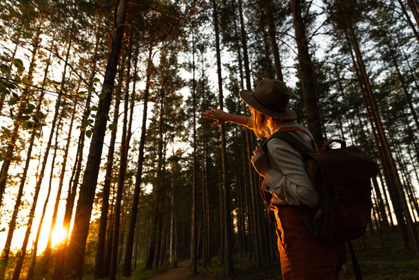 Kobieta przyrodnik w kapeluszu i pomarańczowych ogrodniczkach z plecakiem patrząc na słońce przez palce o zachodzie słońca, odkrywając dziką przyrodę i ekoturystykę przygoda chodzenie w parku narodowym dzikiej przyrody. - Zdjęcie, obraz