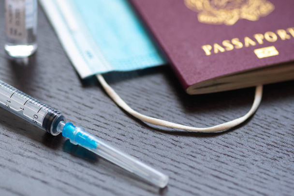 Ruisku neulalla, injektiopullolla, kirurgisella kasvonaamiolla ja passilla tai viisumilla valkoisella pöydällä käyttövalmiina. Covid- tai Coronavirus-rokotteen tausta - Valokuva, kuva