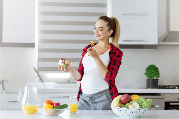 Привлекательная улыбающаяся беременная женщина, держащая миску салата, стоя на кухне, образ жизни дома - Фото, изображение