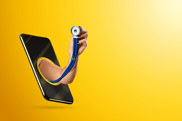 A hand crawling out through a smartphone holds a stethoscope on a yellow background. Online doctor appointment, remote medicine concept. The doctor can examine the patient using a video call - Foto, Bild