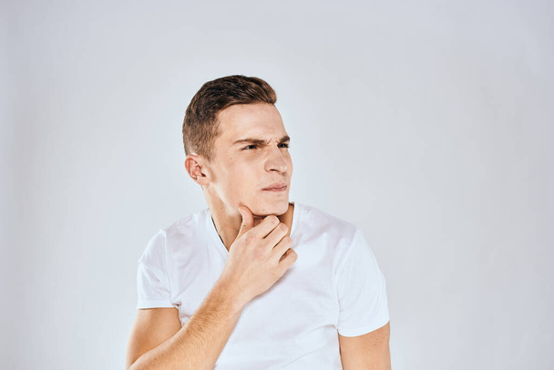 Homem emocional em uma camiseta branca segura a mão em seu rosto fundo claro - Foto, Imagem
