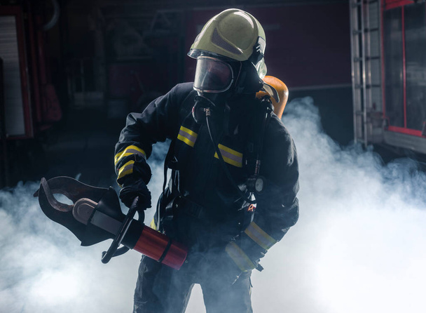 Πορτρέτο πυροσβέστη με πυροσβέστες και κράνος. Σκούρο φόντο με καπνό και μπλε φως. - Φωτογραφία, εικόνα