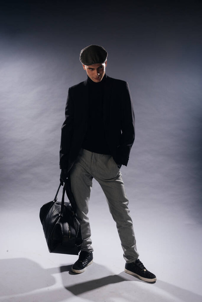 Ritratto di un giovane uomo dalla mascella affilata che indossa un berretto da newsboy, che tiene una borsa da uomo mentre posa in studio su uno sfondo bianco. - Foto, immagini