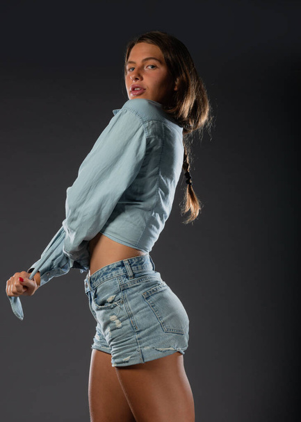 Beautiful young female model in short denim jeans on dark background. - Valokuva, kuva