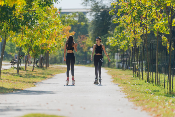 Vue arrière de deux jeunes femmes en forme sur patins à roulettes qui roulent à l'extérieur dans la rue urbaine du parc. - Photo, image