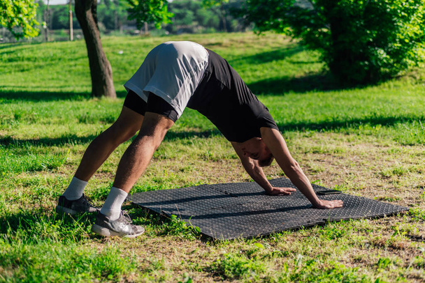 Junger kaukasischer Mann praktiziert Yoga auf einer Matte im Park in nach unten gerichteter Hundepose - Foto, Bild