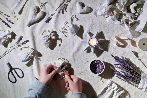 Hand made lavender sachets in textile bags, soft hearts. Hands make sachet. Dry lavender flowers for stuffing, scissors, candle. Flat lay, textile tablecloth. Zero waste, low impact sustainable life. - Valokuva, kuva