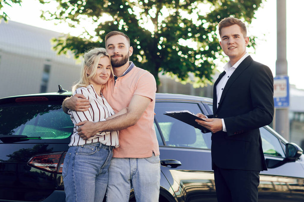 Verkäufer und glückliches junges Paar mit neuem Auto. Junges Paar steht umarmt neben einem Auto im Freien. Autokauf - Foto, Bild
