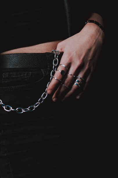 Un primer plano de varios anillos delgados en una mano femenina y una cadena de cintura de cinturón grueso en pantalones negros - Foto, Imagen