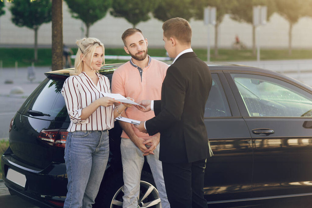 Ένας νεαρός πωλητής δείχνει ένα νέο αυτοκίνητο στους πελάτες. Ευτυχισμένο ζευγάρι, άντρας και γυναίκα αγοράζουν καινούργιο αμάξι. Ο πωλητής δίνει στο νεαρό ζευγάρι έγγραφα για ένα νέο αυτοκίνητο. - Φωτογραφία, εικόνα