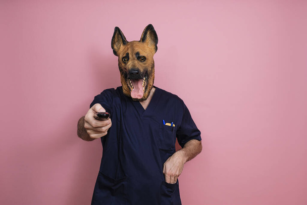 A young man in a latex dog head mask holding a remote control on a pink background - Zdjęcie, obraz
