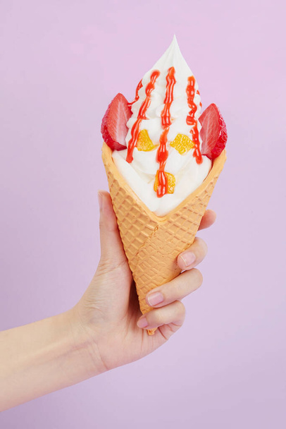 A female's hand holding delicious soft ice cream with fruits and topping in a crispy waffle cone on a purple background - Fotografie, Obrázek