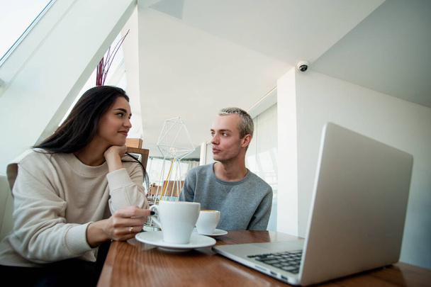 Man and woman looking at each other with interest while spending time in a coffee shop. Blurred modern laptop on front. Spacious light coffee shop on background. Attractive young couple. - Photo, Image