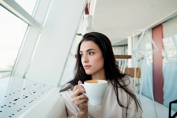 Atraktivní žena s šálkem cappuccino dívá do okna a přemýšlí o něčem. Útulná kavárna atmosféra. Bruneta drží bílý šálek a dívá se jinam. Velký světelný pokoj na pozadí. - Fotografie, Obrázek