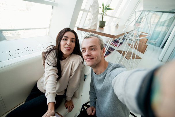 Man and woman taking selfie. Couple having good time in coffee shop. Man holding camera or smartphone when woman pose and showing tongue. Coffee house with panoramic windows on background. - Photo, Image