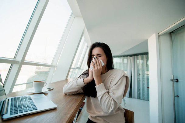 Young woman sneezes in public place. Freelancer working on her modern laptop at coffeehouse blows her nose using paper napkin. Woman got sick due to cold weather outside. Stay home if you are sick - Foto, afbeelding