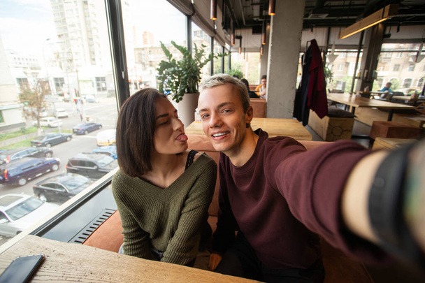 Couple makes crazy faces on camera. Coffee shop on background. Blond man with short hair holds camera and smiling when her girlfriend showing a tongue to him. Stylish man and woman. - Foto, afbeelding
