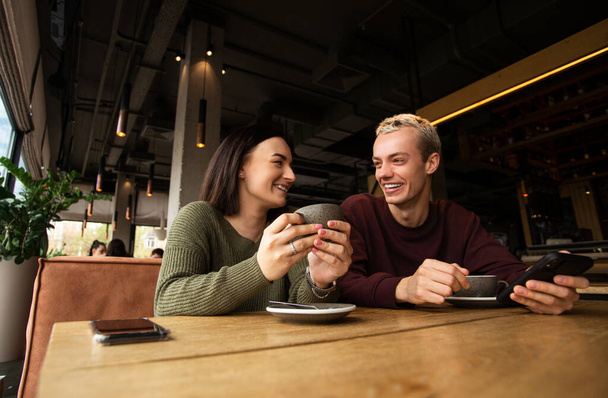 Caucasian couple drinks coffee in cafe. Brunette woman warms her hands on the cup she holds in her hands and smiles to blonde man in purple sweatshirt. Cozy atmosphere in coffee shop. - Foto, Imagem